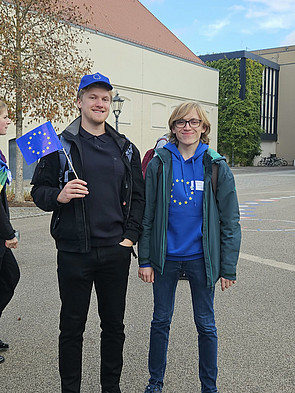 Links steht Felix Brand mit einer EU-Käppi und einer kleinen EU-Fahne in seiner rechten Hand. Rechts steht Linus Sturm, er hat einen EU-Pullover an. Im Hintergrund das Stadthaus Wittenberg.