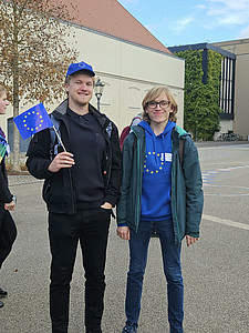 Links steht Felix Brand mit einer EU-Käppi und einer kleinen EU-Fahne in seiner rechten Hand. Rechts steht Linus Sturm, er hat einen EU-Pullover an. Im Hintergrund das Stadthaus Wittenberg.